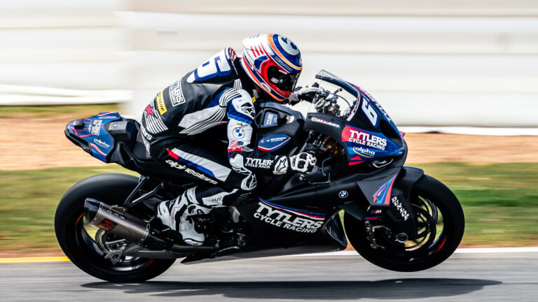 A motorcycle racer in black and white leathers leans forward on a dark race bike at Moto America 2023.