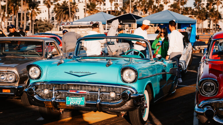 A turquoise vintage convertible with chrome trim is displayed among cars and visitors at the Santa Monica Pier Car Show.